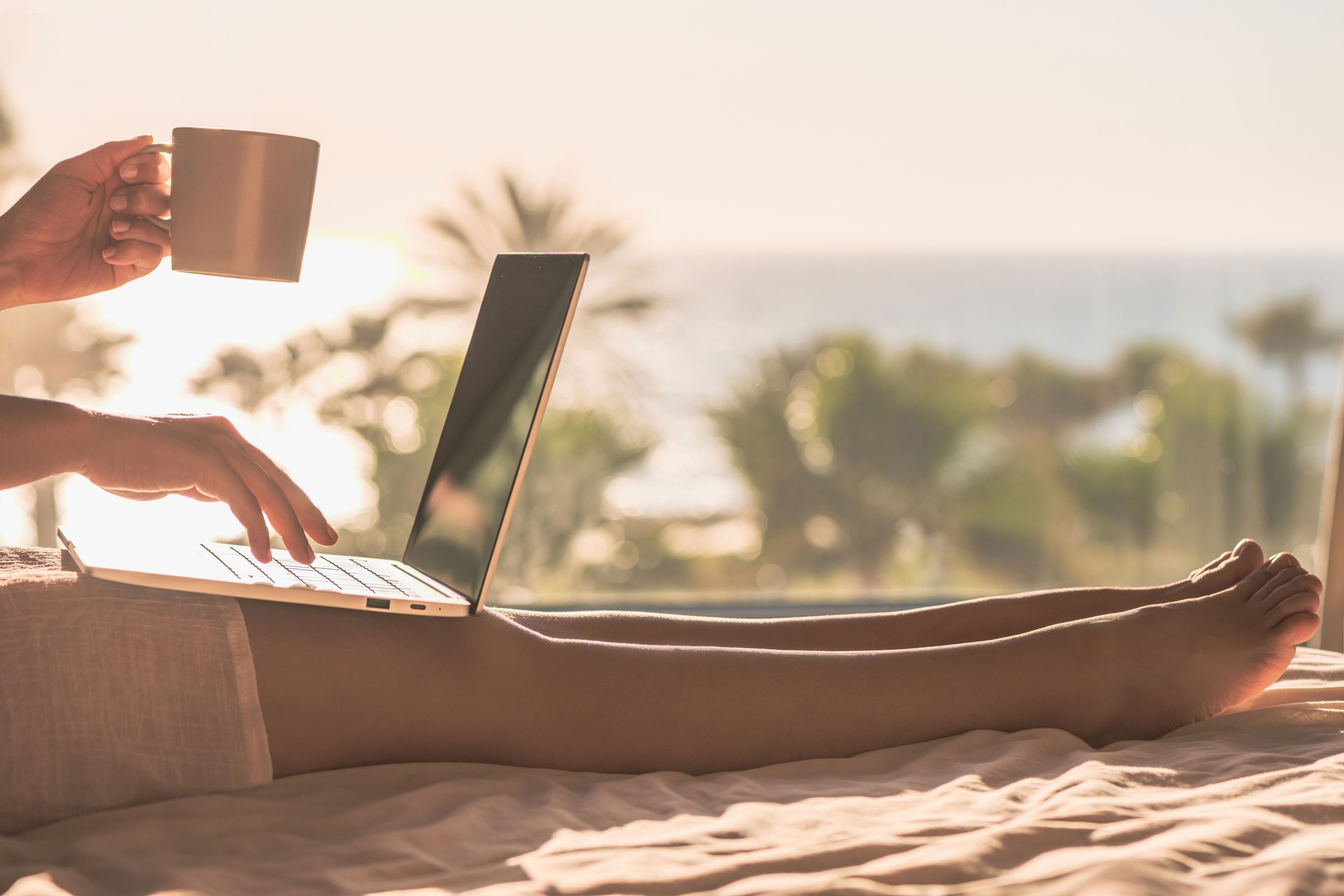 Woman freelancer working with laptop holding cup of coffee sitting on the bed in bedroom near panoramic window with view on palm tree sea beach at sunrise